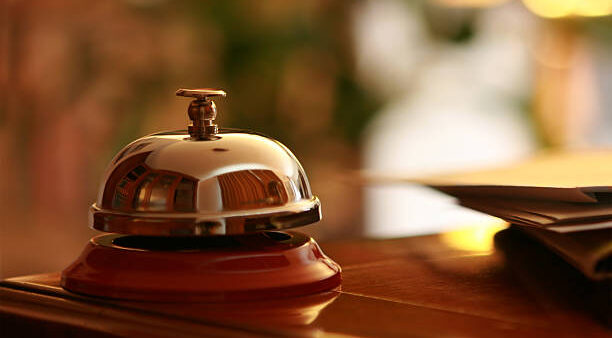 Close-up of a metal and red hotel service bell on a counter of a hotel reception. Business mail laying next to it. Light blur background of a lobby provides space for your text.